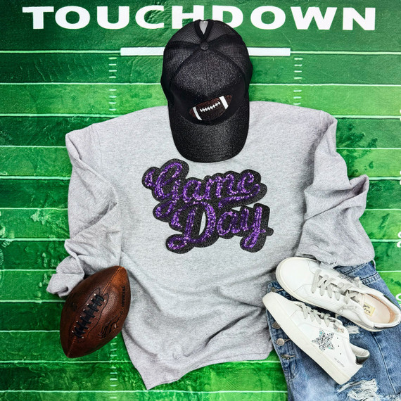 Purple Sequins Game Day Patch, a sparkly purple and black embroidered patch on a grey sweatshirt, displayed on a sports field background with a football, white sneakers, a grey cap with a football embroidery, and a pair of jeans arranged around it.