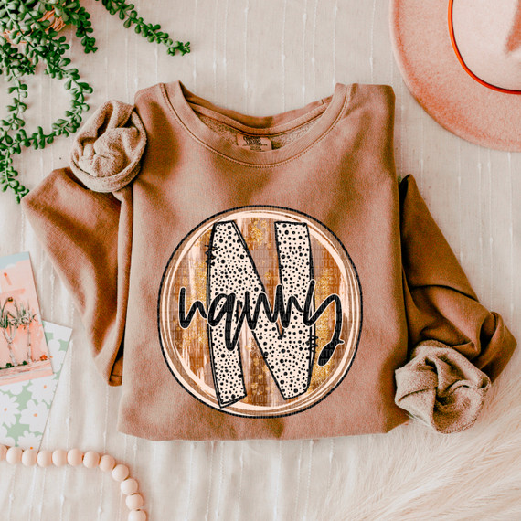 Nanny Fall Brushstrokes DTF Heat Transfer, an image of a heat transfer design on a beige sweatshirt featuring a large letter 'N' with a brushstroke pattern and the word 'nanny' written across it.