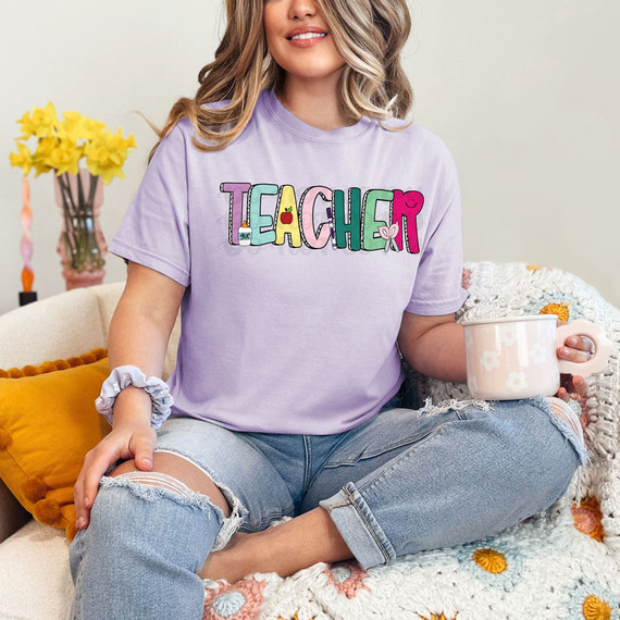 Teacher Dots DTF Heat Transfer, colorful and playful design on a purple T-shirt worn by a woman sitting on a couch with a mug in her hand.