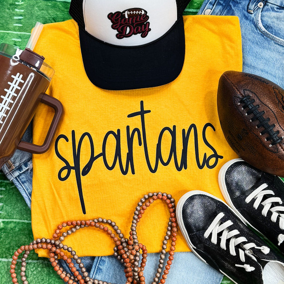Spartans Black PUFF Screen Print Heat Transfer, black and orange text design on a yellow fabric shirt surrounded by sports accessories.