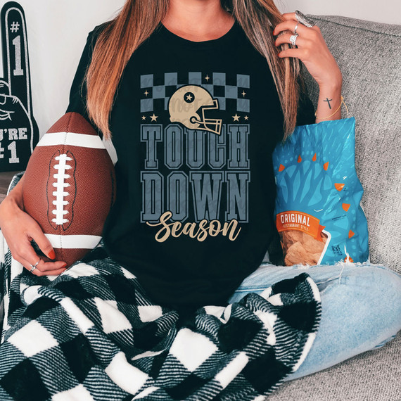 Touchdown Season DTF Heat Transfer, a person sitting on a couch holding a football and a bag of chips, wearing a black T-shirt with football-themed design and text.