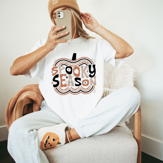 Spooky Season Pumpkin DTF Heat Transfer, woman wearing a white T-shirt with Halloween-themed design, sitting on a cozy chair, taking a selfie with a smartphone.