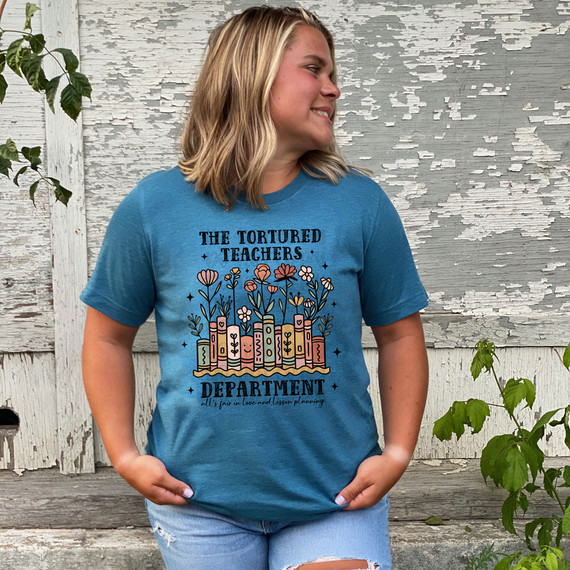 The Tortured Teachers Department DTF Heat Transfer, a colorful design featuring books, flowers, and text, printed on a blue t-shirt worn by a woman.