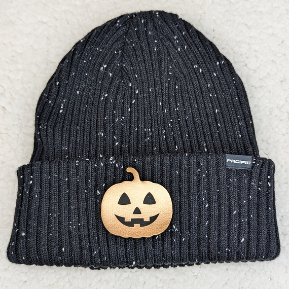Jack O Lantern Faux Leather Patch, a decorative sewn patch with a smiling pumpkin face, placed on a black knit beanie with specks of white.