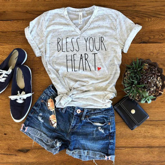 Bless your heart sublimation transfer, casual outfit with graphic T-shirt, denim shorts, sneakers, sunglasses, wallet, and succulent plant on wooden background.
