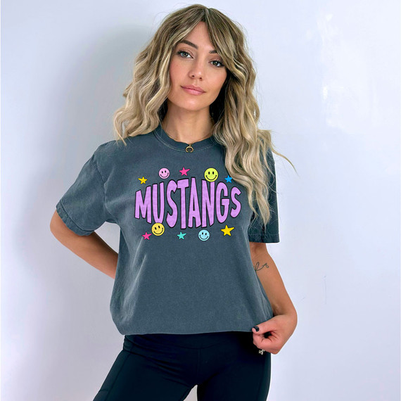 Mustangs Happy Face DTF Heat Transfer, colorful graphic design on a woman's T-shirt with smiling faces, stars, and the word 'MUSTANGS' in purple.