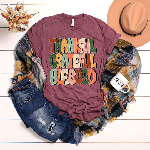Thankful Grateful Blessed Retro DTF Heat Transfer T-shirt with colorful lettering on a maroon fabric, accompanied by denim shorts, a plaid scarf, a ceramic mug, and a beige hat on a white wooden background.
