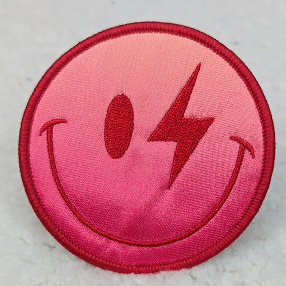 Red Ombre Smile Embroidered HAT/POCKET Patch, circular patch with a pink background, red embroidered smiley face, lightning bolt nose, and curved smile line.