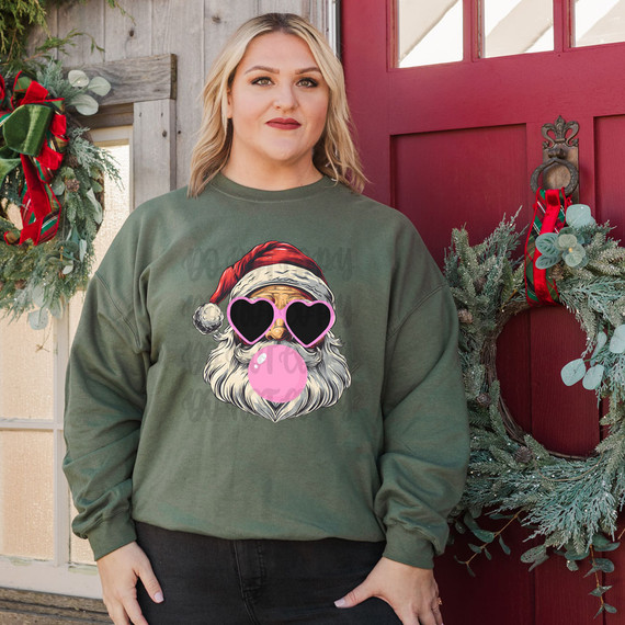 Santa Blowing Bubblegum DTF Heat Transfer, humorous holiday graphic featuring Santa with heart-shaped sunglasses and blowing pink bubblegum, on a woman's green sweatshirt for Christmas festivities.