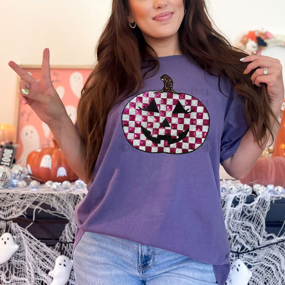 FAUX Sequin Checkered Pumpkin DTF Heat Transfer, a close-up of a woman wearing a purple t-shirt with a checkered sequin pumpkin face design, surrounded by Halloween decor.