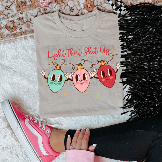 Light That Shit Up DTF Heat Transfer, colorful cartoon Christmas light bulbs with cute faces and hand gestures on a beige t-shirt.