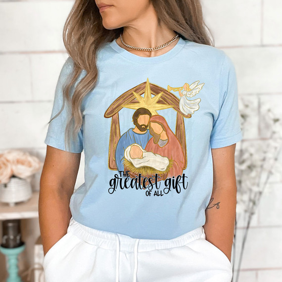 The Greatest Gift Of All DTF Heat Transfer, a woman wearing a light blue t-shirt with a festive nativity scene design of Mary, Joseph, baby Jesus, and an angel.