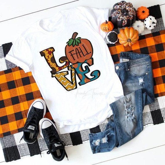 Love Fall Sublimation Transfer, festive fall-themed design on a white t-shirt featuring colorful autumn elements and text, surrounded by small decorative pumpkins and plaid patterned cloth with jeans and sneakers.