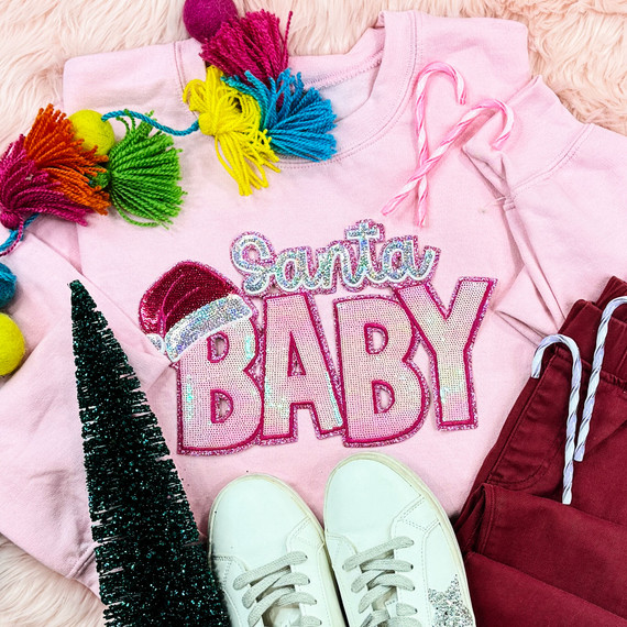 Santa Baby With Hat Sequin Patch, pink sweatshirt with 'Santa Baby' text and Santa hat sequin patch, surrounded by colorful pom-poms, candy canes, a mini Christmas tree, and white sneakers.