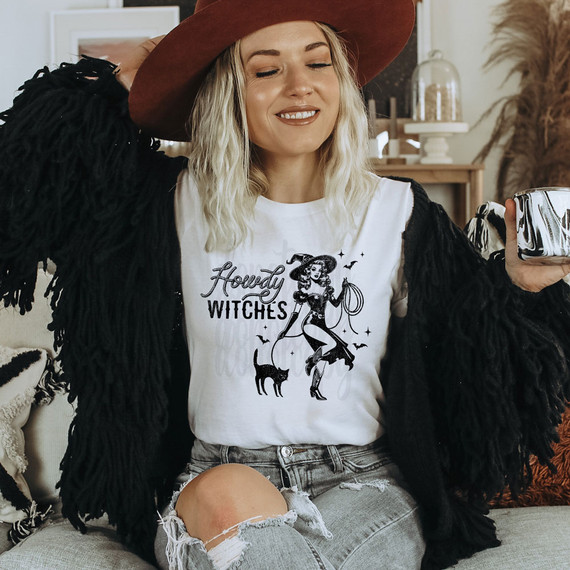 Howdy Witches Pin Up DTF Heat Transfer, woman wearing a white t-shirt with a witch illustration and text, wearing a wide-brimmed hat and a black furry coat.
