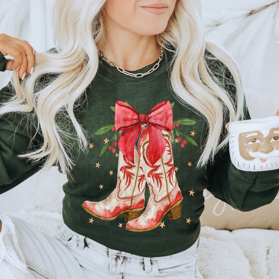 Christmas Cowgirl Boots DTF Heat Transfer, festive Christmas-themed design on a woman's sweatshirt featuring cowboy boots with a red bow, holly leaves, and berries.