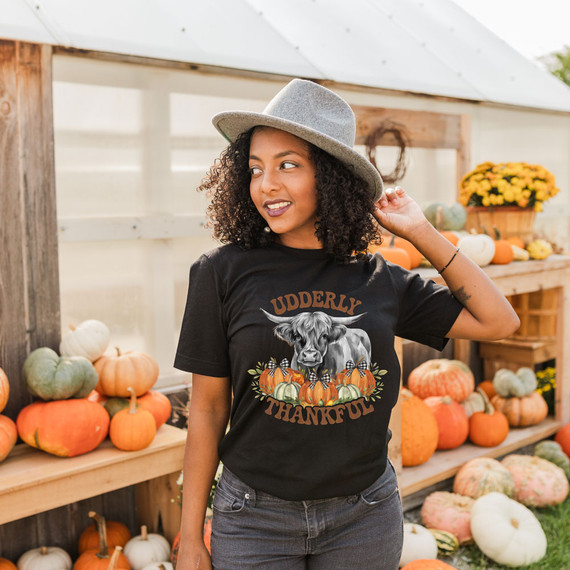 Udderly Thankful DTF Heat Transfer, woman wearing black T-shirt with autumn pumpkin and cow graphic in a pumpkin patch setting.