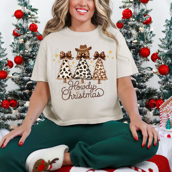 Howdy Christmas DTF Heat Transfer, festive woman wearing holiday-themed graphic T-shirt featuring stylized Christmas trees with animal print patterns and bows, sitting in front of decorated Christmas trees with red ornaments.
