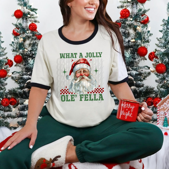 What A Jolly Ole' Fella DTF Heat Transfer, person wearing Christmas-themed shirt with Santa Claus graphic and holiday message, sitting by decorated Christmas trees.