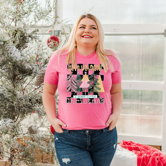 Have Yourself A Merry Little Christmas Trees DTF Heat Transfer, a woman wearing a pink T-shirt with Christmas tree designs and holiday-themed text, standing next to a decorated Christmas tree.
