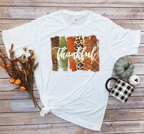 Thankful word art fall colors plaid leopard glitter pattern sublimation transfer, on a white T-shirt with fall-themed decorations around, including dried flowers and pumpkins.