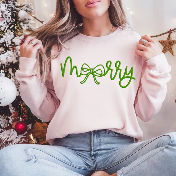 Merry Bow Green PUFF Screen Print Heat Transfer, a green holiday design with a bow incorporated into the lettering, on a pink sweatshirt worn by a woman in a festive setting.