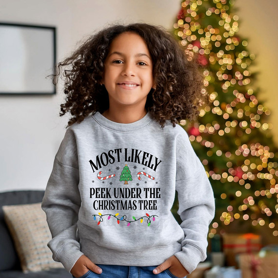 Most Likely Peek Under The Christmas Tree Festive DTF Heat Transfer, Christmas-themed design with a decorated tree, candy canes, and colorful lights on a sweatshirt worn by a smiling girl with curly hair in front of a Christmas tree.