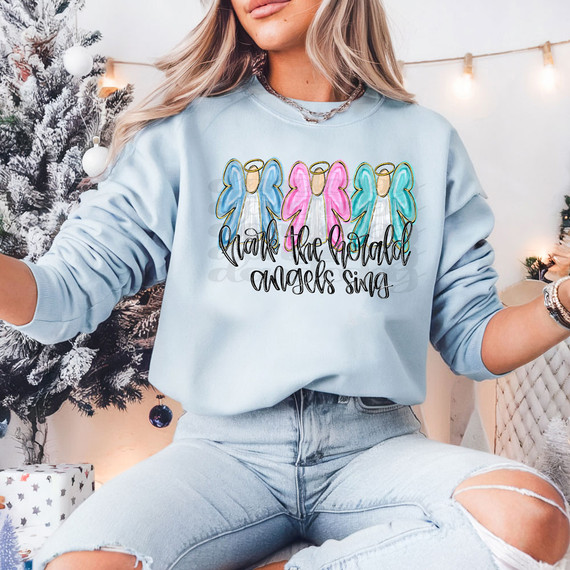 Hark The Heralds Angels Sing DTF Heat Transfer, Christmas-themed sweatshirt with angel designs and festive message, worn by a woman near decorated Christmas tree and holiday lights.