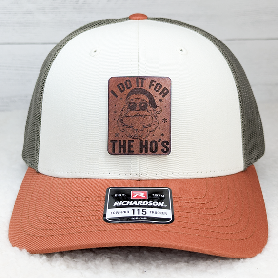 I Do It For The Ho's Leather Hat Patch, leather patch with Santa's face wearing sunglasses and a beard, with the text I Do It For The Ho's, on a trucker hat featuring a white front panel, grey mesh back, and rust-colored brim.