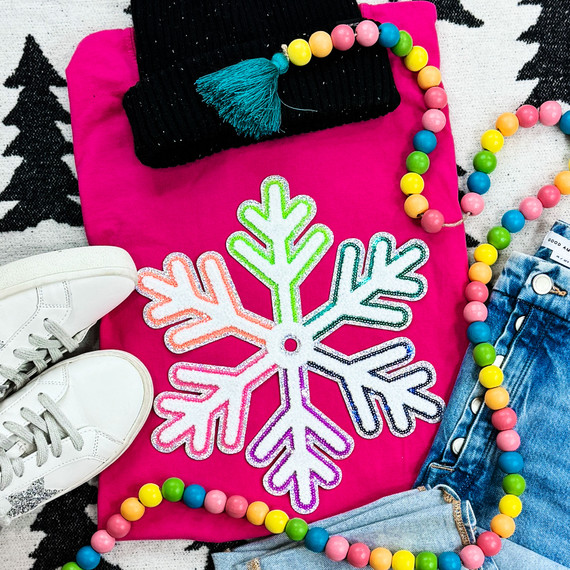 Multi-Colored Chenille/Sequin Snowflake Patch, vibrant snowflake patch with multi-colored edges on a pink background surrounded by white sneakers, a black beanie with a blue tassel, a colorful bead necklace, and denim jeans.