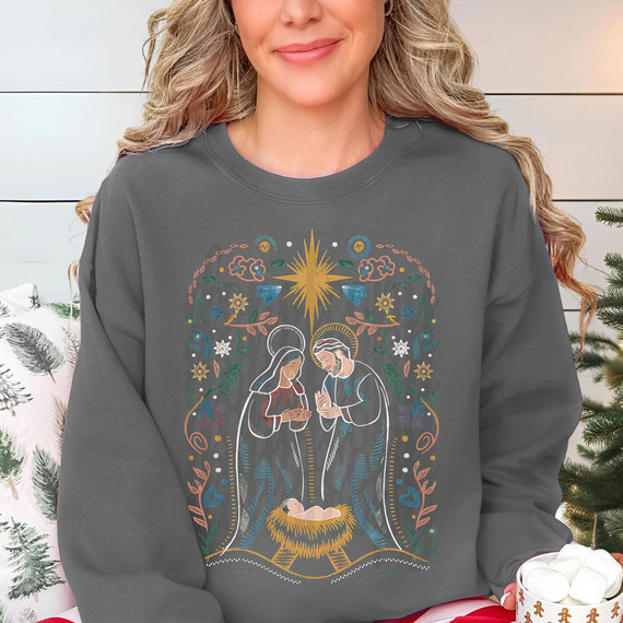 Vintage Nativity DTF Heat Transfer, woman wearing a Christmas-themed sweatshirt featuring a stylized nativity scene with Mary, Joseph, Baby Jesus, and decorative elements in vibrant colors.