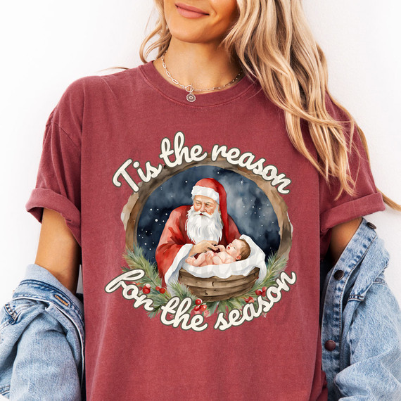 Tis The Reason For The Season DTF Heat Transfer, Christmas-themed graphic of Santa Claus holding a baby Jesus in a manger surrounded by holly and pine branches.