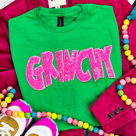 Grinchy Pink Sequin Patch, vibrant green sweatshirt with pink glittery 'GRINCH' patch, surrounded by colorful beads, pink shoes, and pink textured fabric.