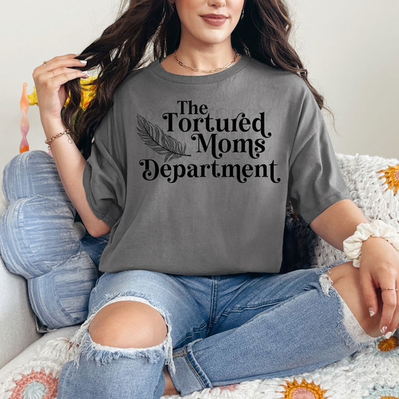 The Tortured Moms Department DTF Heat Transfer, a black text and feather design printed on a gray T-shirt worn by a woman sitting on a cozy sofa.