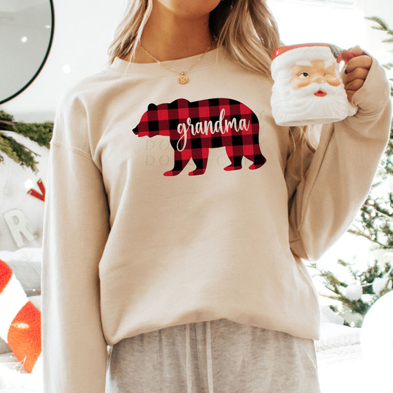 Grandma Bear Red Buffalo Plaid DTF Heat Transfer, close-up of a woman wearing a beige sweatshirt featuring a bear silhouette in red buffalo plaid pattern with the word grandma written inside, holding a Santa mug, Christmas decorations in the background.