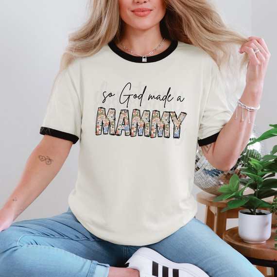 So God Made A Mammy DTF Heat Transfer, woman wearing a cream T-shirt with black collar and cuffs, featuring the text 'So God Made A Mammy' in a floral-patterned font, in a home decor setting.