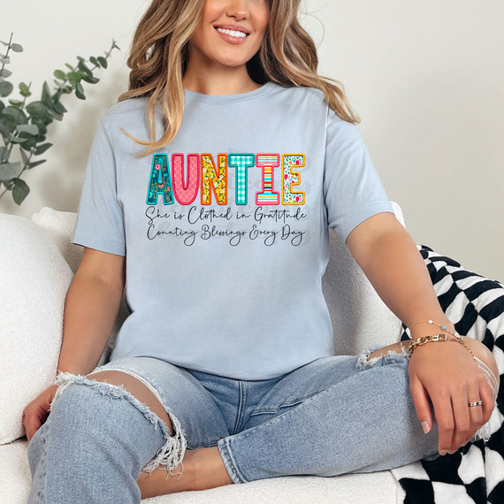 Auntie She Is Clothed In Gratitude Counting Blessings Every Day DTF Heat Transfer, a woman wearing a light blue t-shirt with colorful embroidered alphabet spelling 'AUNTIE' and a heartfelt message beneath.