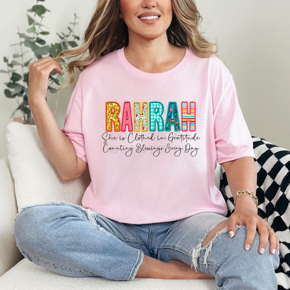 RahRah She Is Clothed In Gratitude Counting Blessings Every Day DTF Heat Transfer, woman wearing a pink T-shirt with colorful and patterned text, sitting on a white couch with a plant in the background.