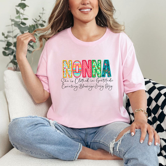 Nonna She Is Clothed In Gratitude Counting Blessings Every Day DTF Heat Transfer, woman wearing pink T-shirt with colorful 'NONNA' text and gratitude message, casual sitting on a sofa.
