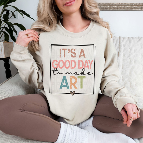 It's A Good Day To Make Art DTF Heat Transfer, a woman wearing a cream sweatshirt with colorful text and design, sitting on a sofa in a cozy room.