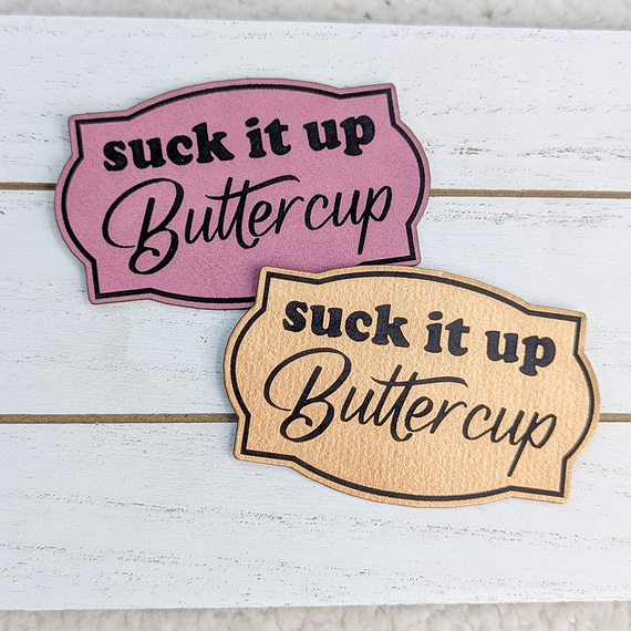 Suck It Up Buttercup FAUX Leather Patch, pink and tan faux leather patches with the phrase "suck it up Buttercup" in black text on a white wooden background.