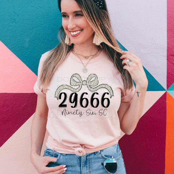 Ninety Six, SC Zip Code Bow DTF Heat Transfer, customized t-shirt design with number 29666 and text Ninety Six, SC, worn by a smiling woman against a colorful mural background.
