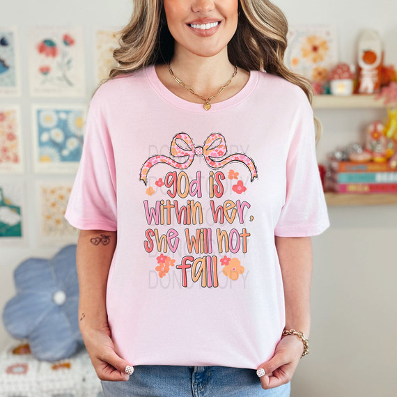 God Is Within Her She Will Not Fall DTF Heat Transfer, woman wearing a pink t-shirt with inspirational quote and decorative bow design, in a colorful craft room setting.
