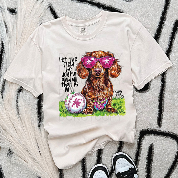 Let The Field Be Joyful And All That Is In It DTF Heat Transfer, white t-shirt with a graphic of a dog wearing pink sunglasses and a colorful bow, sitting on green grass near a baseball with pink flower design.