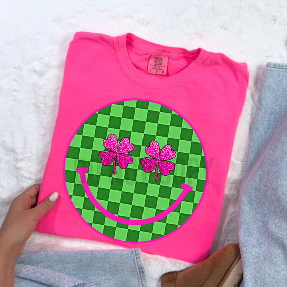 Checkered Shamrock Happy Face DTF Heat Transfer, pink t-shirt with a green checkered background featuring pink glittery shamrock and happy face design.