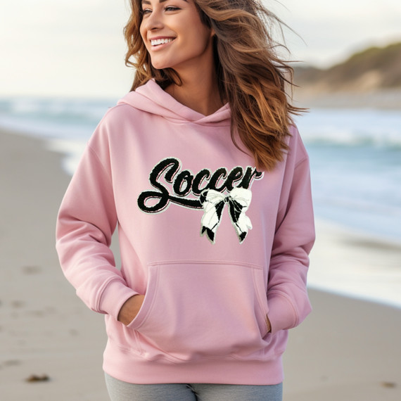 Soccer With Bow Chenille Patch, women wearing a pink hoodie with a soccer-themed chenille patch of a bow and the word 'Soccer' on the beach.