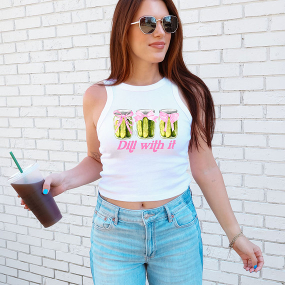Dill With It Coquette DTF Heat Transfer, a woman wearing a white sleeveless top with a colorful graphic of three jars with pink bows and the text 'Dill with it' in pink, standing in front of a white brick wall.