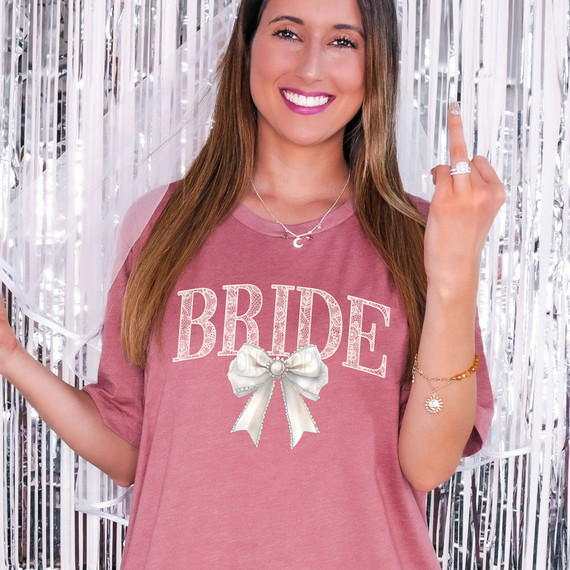 Bride Faux Lace Coquette DTF Heat Transfer, a woman showing a customized T-shirt with the word 'BRIDE' and a satin bow design for bridal events.
