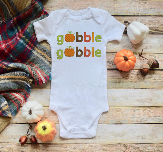 Patchwork Gobble Gobble Pumpkin Sublimation Transfer, a cute holiday-themed baby onesie with fall pumpkin graphics and text, laid on a wooden surface next to mini pumpkins and a colorful plaid fabric.