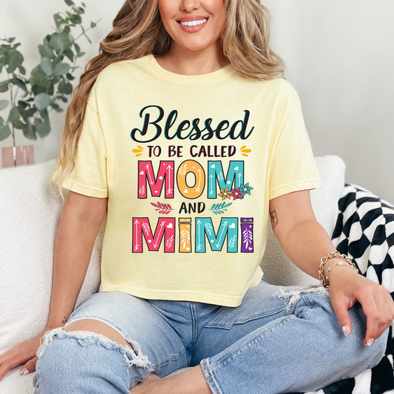 Blessed To Be Called Mom And Mimi DTF Heat Transfer, colorful floral and text design on a light yellow t-shirt worn by a smiling woman sitting on a sofa.